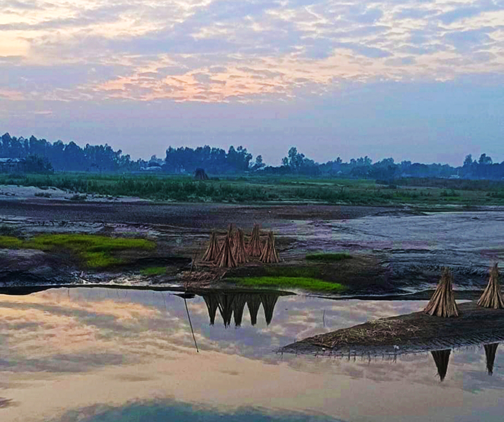 পুরাতন ধলেশ্বরী নদীর পাড়ে শুকানোর জন্য রেখে দেওয়া হয়েছে পাট কাঠি। দূরে বাড়ি-ঘর আর গাছপালার আভাস। সব কিছু মিলিয়ে গোধূলী বেলায় পড়ন্ত সূর্যের আলোয় জন্ম নিয়েছে অপরূপ এক দৃশ্যের। ঘিওরের শ্রীধর নগর এলাকা, মানিকগঞ্জ, ৭ ডিসেম্বর ২০২৩। ছবি: আব্দুর রাজ্জাক